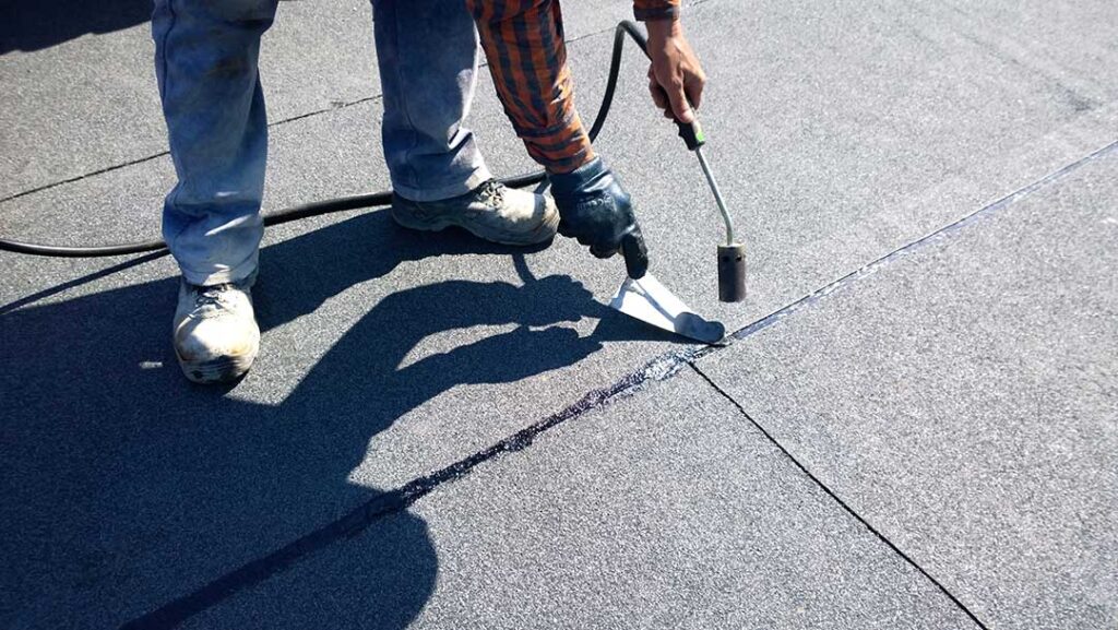 A person applying roofing sealant around a vent pipe on a residential shingle roof.