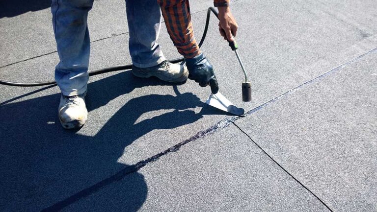 A person applying roofing sealant around a vent pipe on a residential shingle roof.